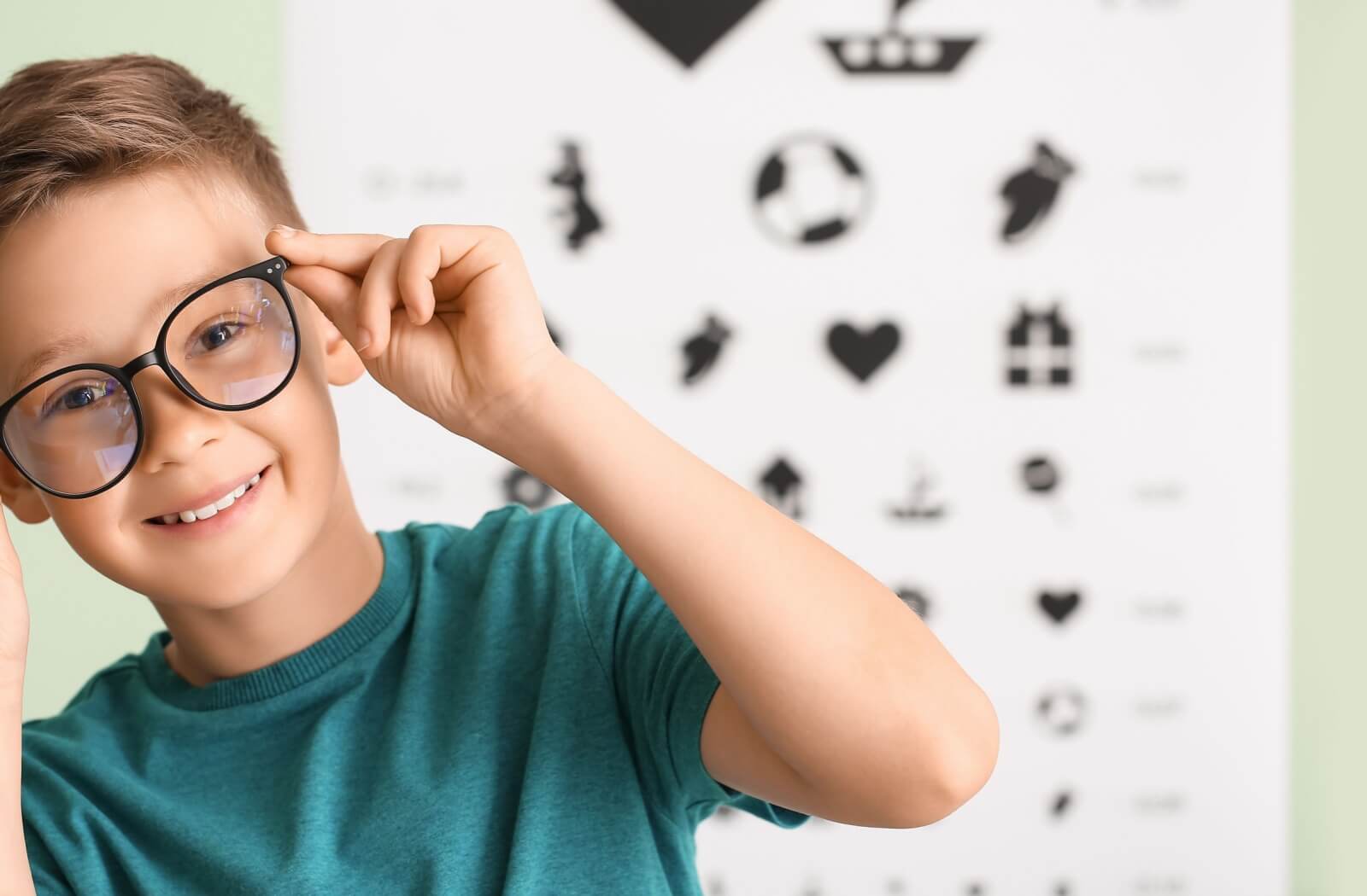 A child in a teal shirt smiling while adjusting a pair of black-rimmed eyeglasses in front of a blurred pediatric eye chart, illustrating the use of myopia eyeglasses.