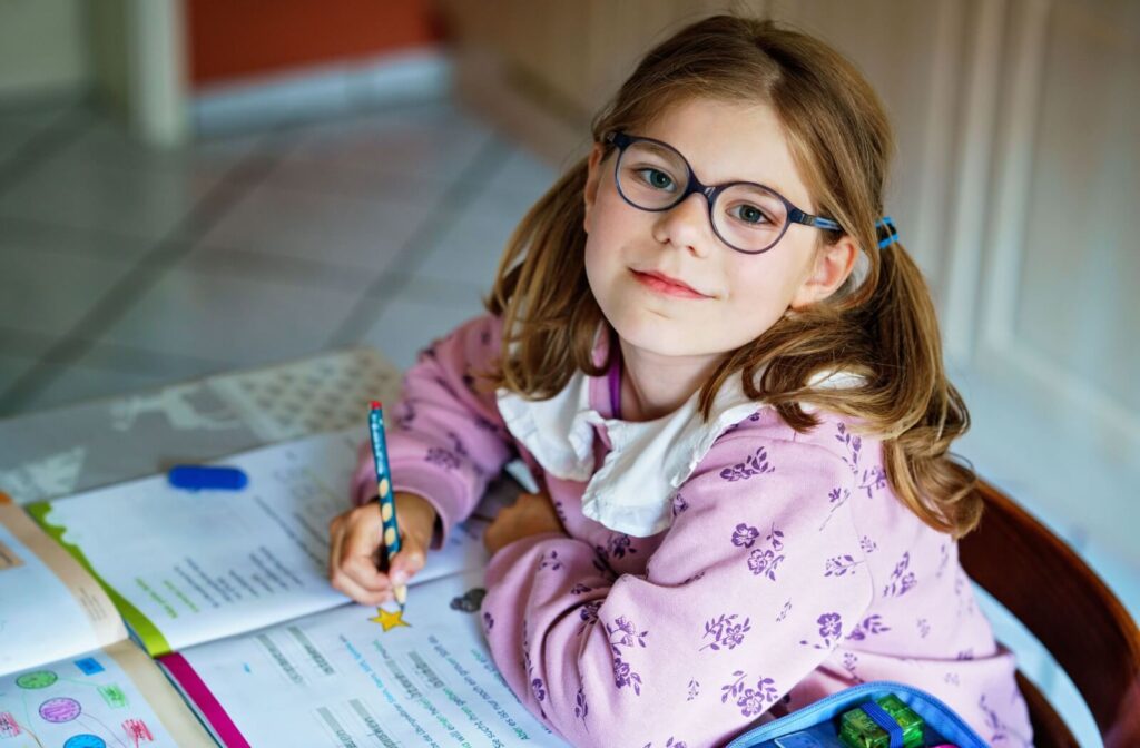 A young person with brown hair in pigtails wearing blue-rimmed eyeglasses while sitting at a desk and writing in a workbook, showcasing the use of myopia eyeglasses for schoolwork.